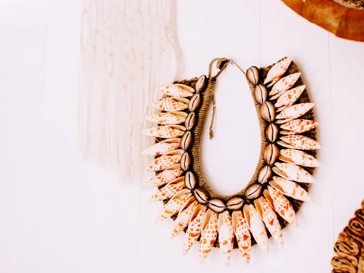 Un grand collier ornemental de coquillages tachetés et de cauris, monté sur fibre, accroché à un mur clair.