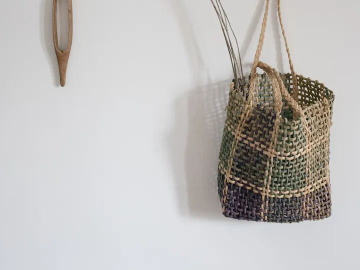 Un cabas ajouré aux fibres teintes, anses tressées, suspendu à un mur clair près de fleurs séchées.